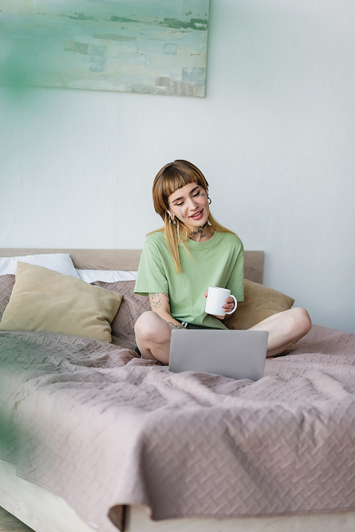 smiling tattooed woman sitting on bed with cup of tea and watching movie on computer