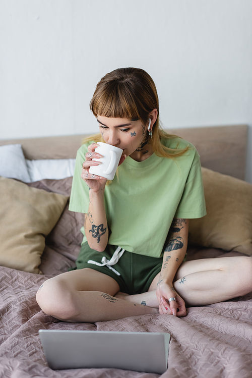 young worried woman drinking tea while watching film on computer in bedroom
