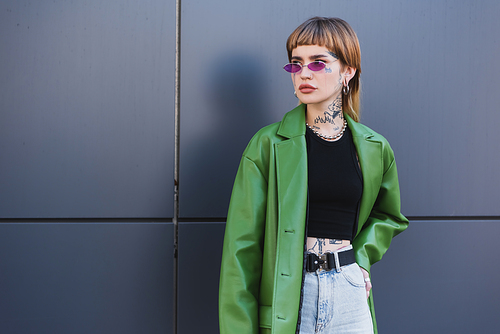 tattooed woman in green jacket standing with hand on hip and looking away near grey wall
