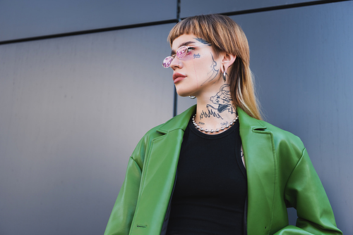 low angle view of tattooed woman in stylish eyeglasses and green jacket looking away near grey wall