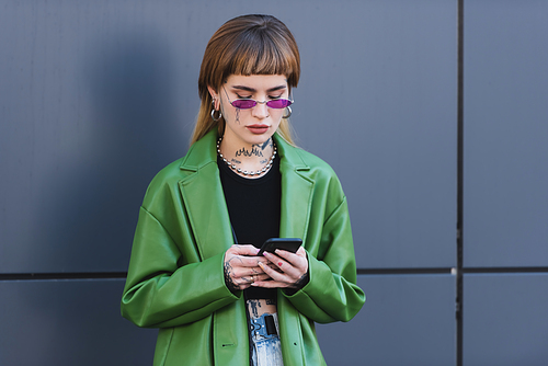 trendy tattooed woman messaging on smartphone near grey wall outdoors