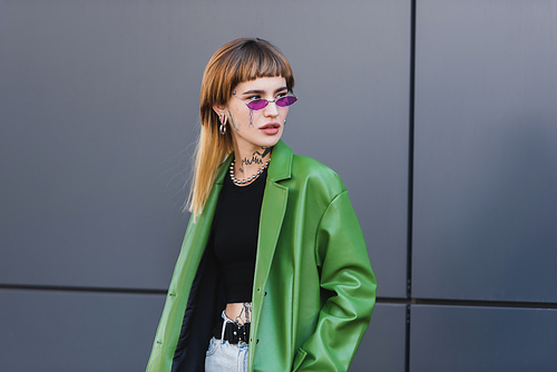 tattooed woman in green leather jacket and trendy eyeglasses looking away on city street