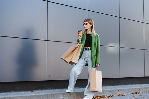 stylish tattooed woman in green jacket and jeans standing with shopping bags and paper cup outdoors
