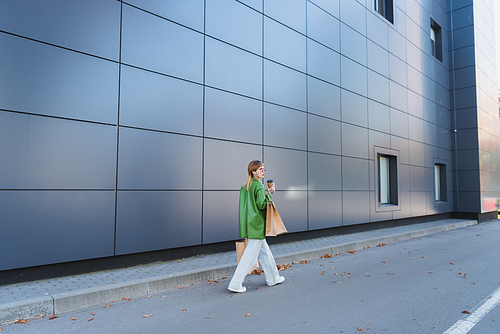 young stylish woman with shopping bags and takeaway drink walking along grey wall