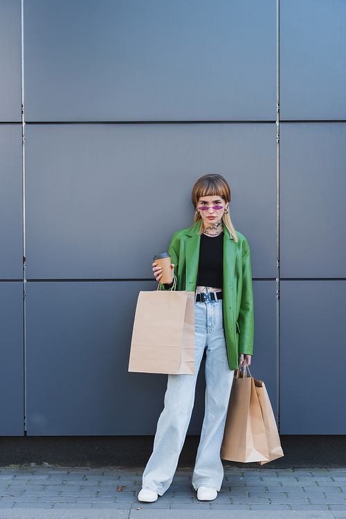 trendy tattooed woman with shopping bags and coffee to go  near grey wall