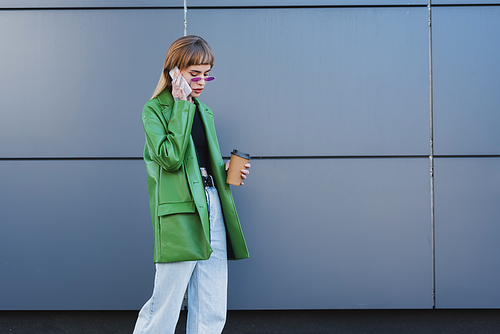 trendy woman in green leather jacket holding paper cup while talking on mobile phone outdoors