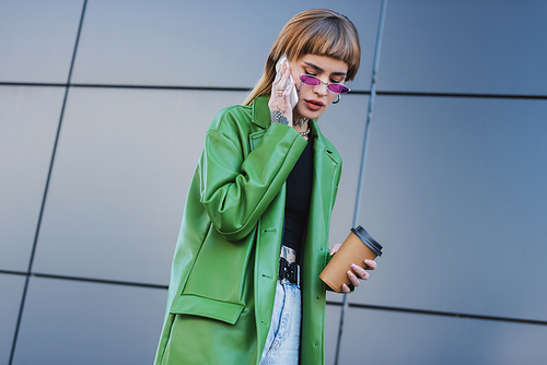 tattooed woman in green leather jacket holding coffee to go and calling on cellphone outdoors