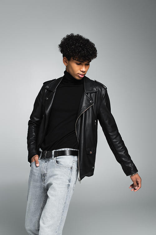 african american man with trendy hairstyle, in leather jacket, posing with hand in pocket on grey