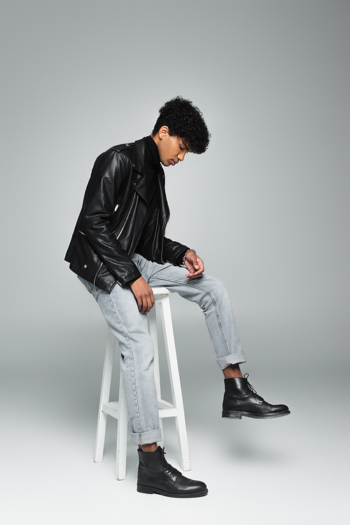 young african american man in jeans, black leather boots and jacket sitting on high stool on grey background