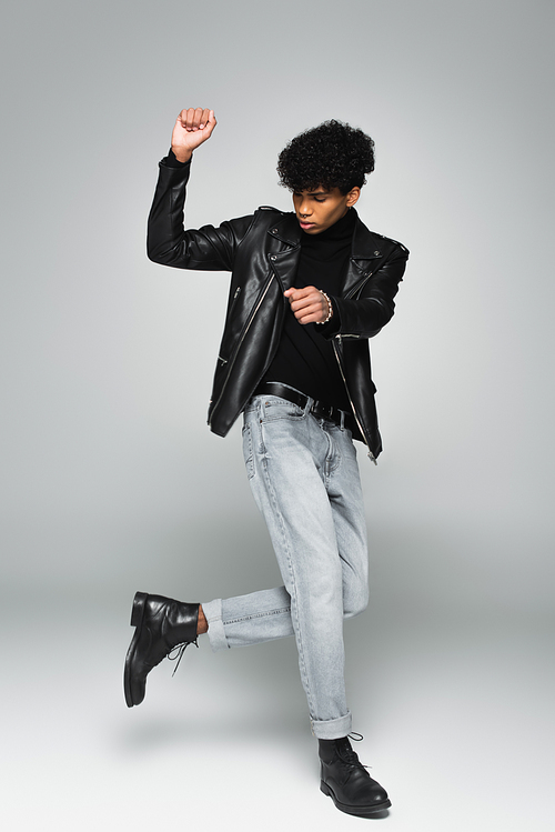 african american man in jeans, black leather jacket and boots dancing on grey background