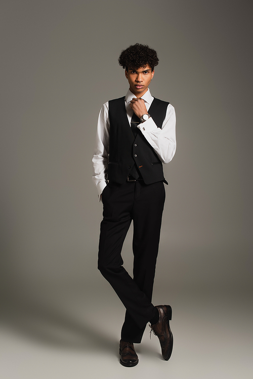 elegant african american man adjusting black tie while posing with hand in pocket on grey background
