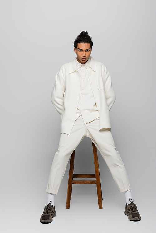 full length view of african american man in white suit and sneakers posing near high stool on grey background