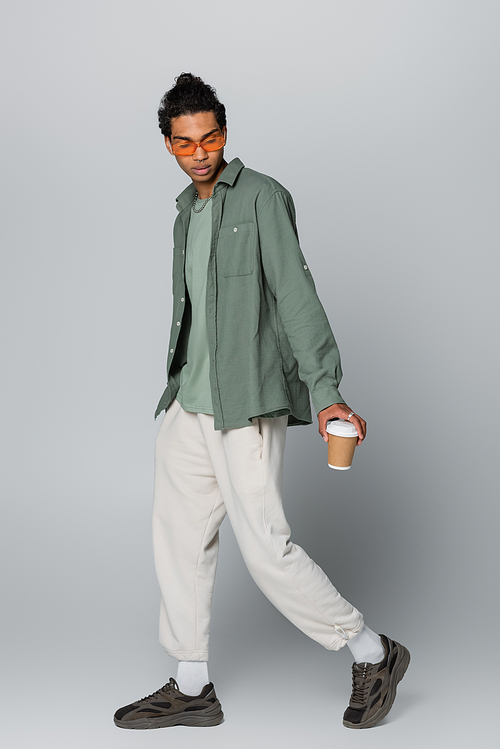 full length view of stylish african american guy walking with coffee to go on grey background