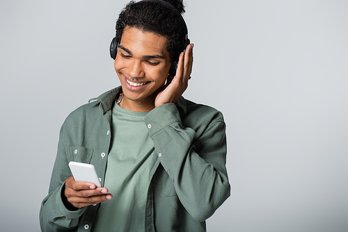 stylish african american guy looking at smartphone while listening music in wireless headphones isolated on grey