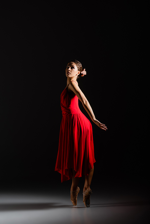 Young ballerina in red dress and pointe shoes dancing on black background