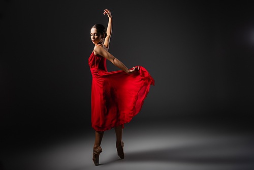 Pretty ballerina holding red dress while dancing on black background