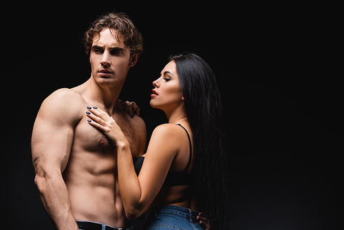 young woman in bra and jeans touching muscular boyfriend isolated on black
