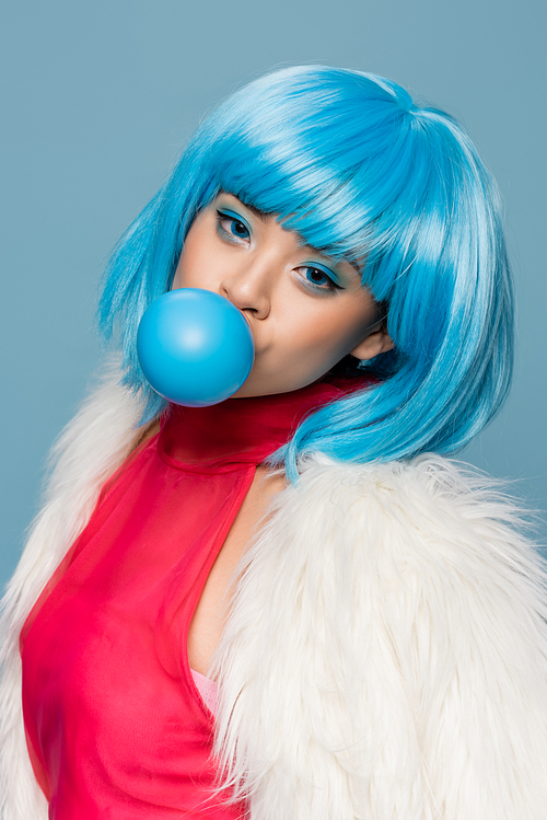 Stylish asian woman in furry jacket and wig blowing bubble gum isolated on blue