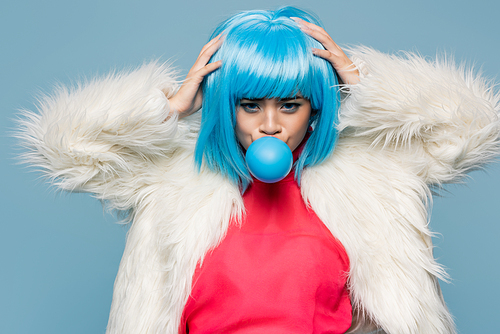 Young asian pop art woman touching wig while blowing bubble gum isolated on blue