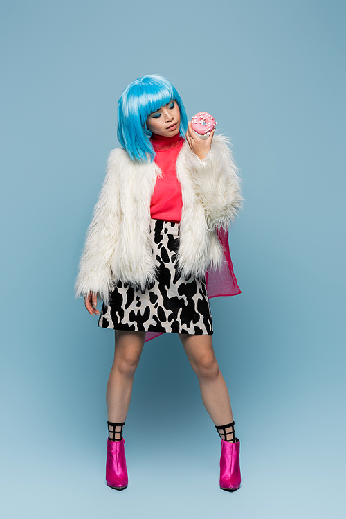 Stylish asian woman in furry jacket looking at donut on blue background