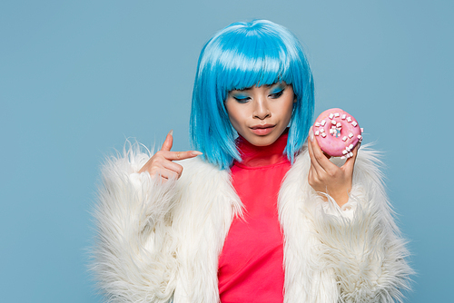 Stylish asian woman in fluffy jacket pointing at donut isolated on blue