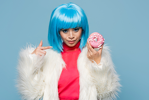 Smiling asian woman in pop art style pointing at donut isolated on blue