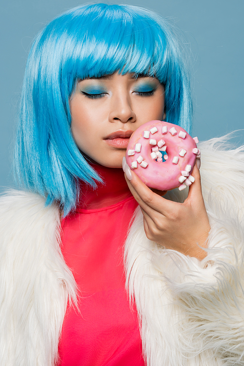 Asian woman in pop art style closing eyes while holding tasty donut isolated on blue