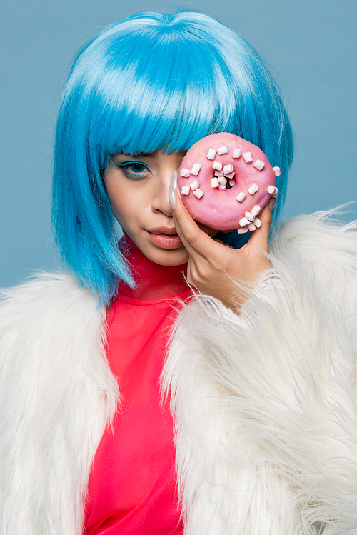 Young asian woman in fluffy jacket and wig holding donut with glaze near face isolated on blue