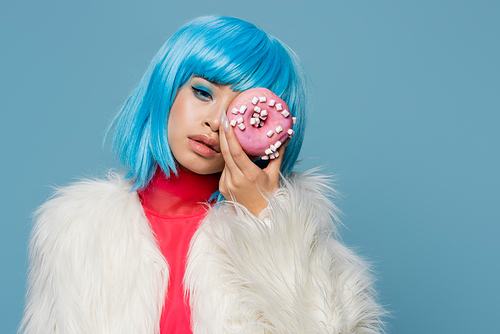 Trendy young asian woman in fluffy jacket holding donut near face isolated on blue