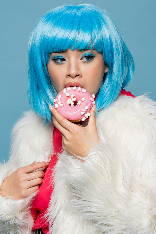 Pretty asian woman in pop art style eating donut with glaze isolated on blue