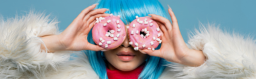 Young woman in wig and fluffy jacket holding donuts near eyes isolated on blue, banner