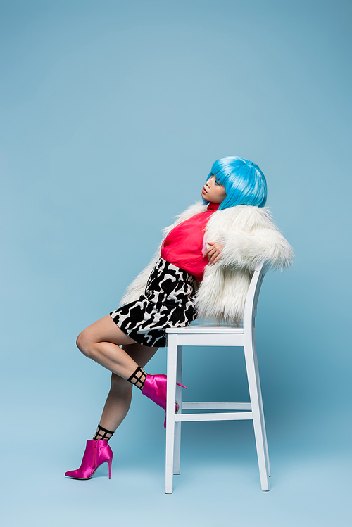 Fashionable woman in fluffy jacket and bright wig posing near chair on blue background