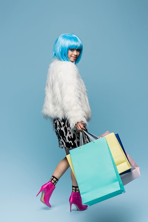 Stylish asian woman in wig and fluffy jacket holding shopping bags and smiling at camera on blue background