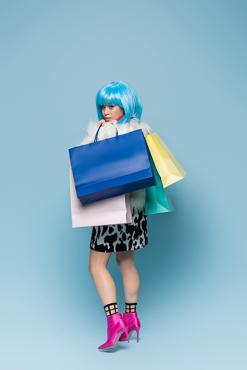 Trendy asian woman in heels and fluffy jacket holding shopping bags on blue background