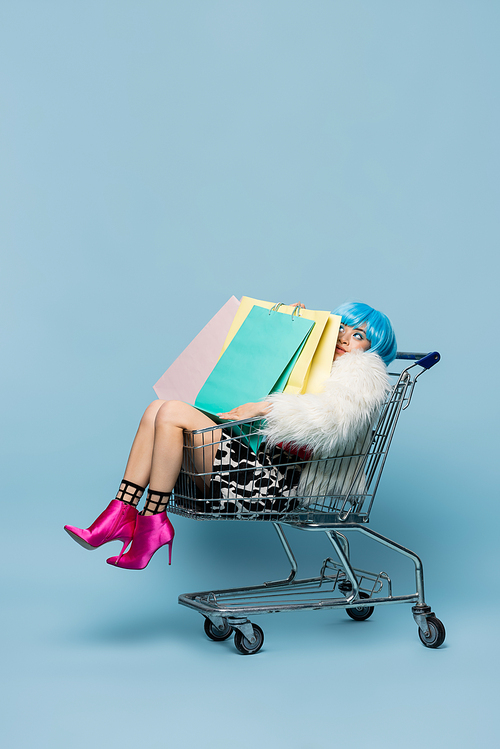 Young asian woman in pop art style sitting in shopping cart with bags on blue background