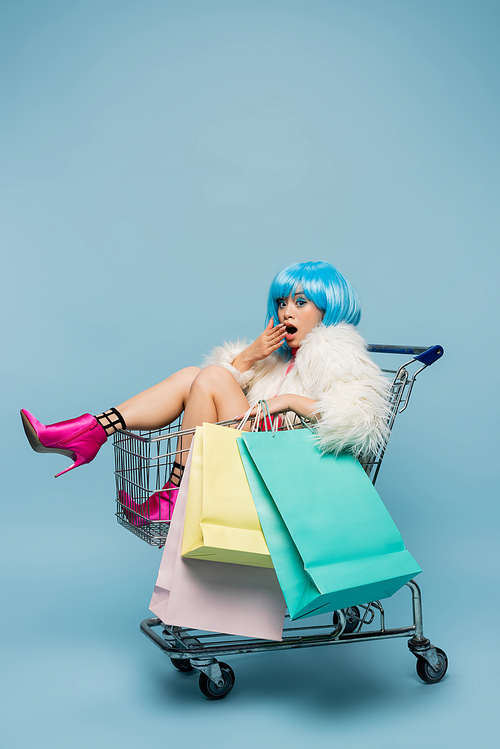 Amazed asian woman in pop art style  while holding shopping bags in cart on blue background