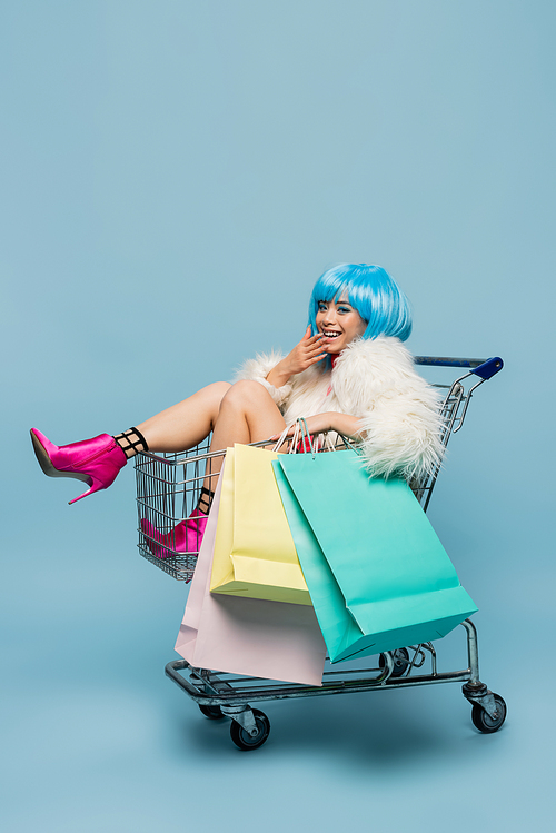 Positive asian pop art woman holding shopping bags in cart on blue background