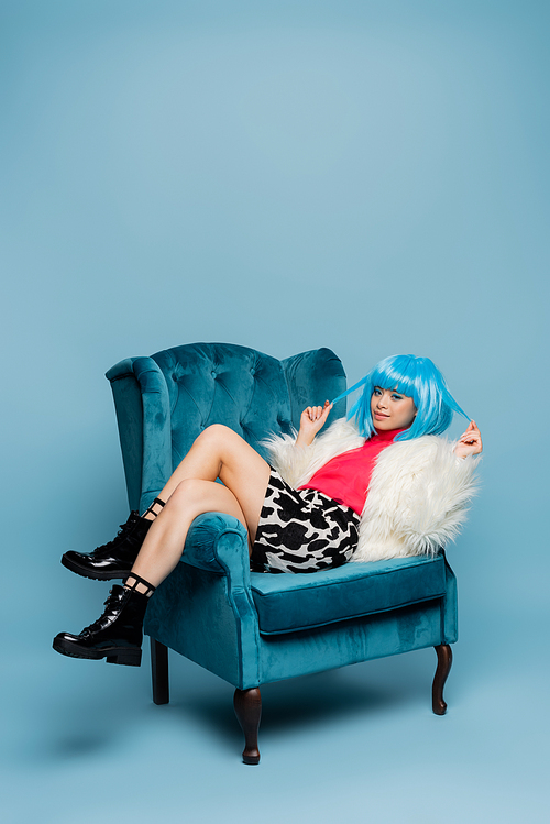 Smiling asian pop art woman touching hair of bright wig in armchair on blue background