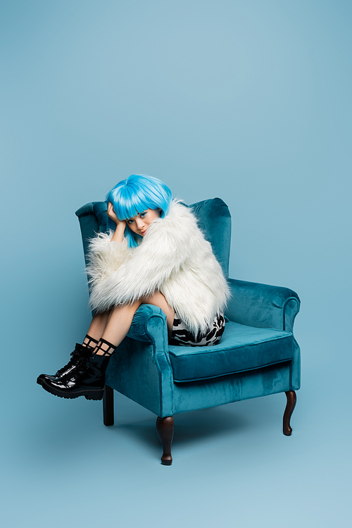 Stylish asian woman in fluffy jacket and wig sitting on armchair on blue background