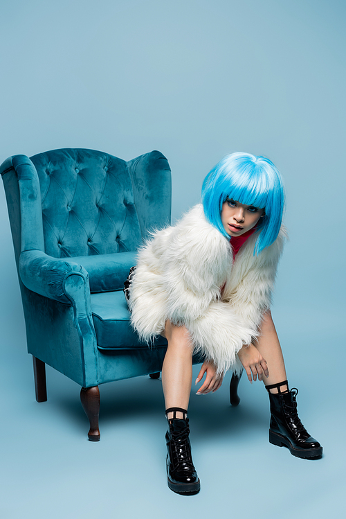 Asian woman in bright wig and fluffy jacket  on turquoise armchair on blue background