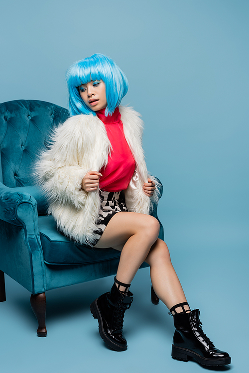 Pretty asian pop art woman in fluffy jacket sitting on velveteen armchair on blue background