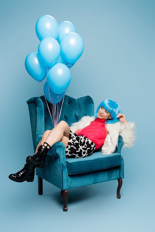 Happy asian model in pop art style looking at balloons on armchair on blue background