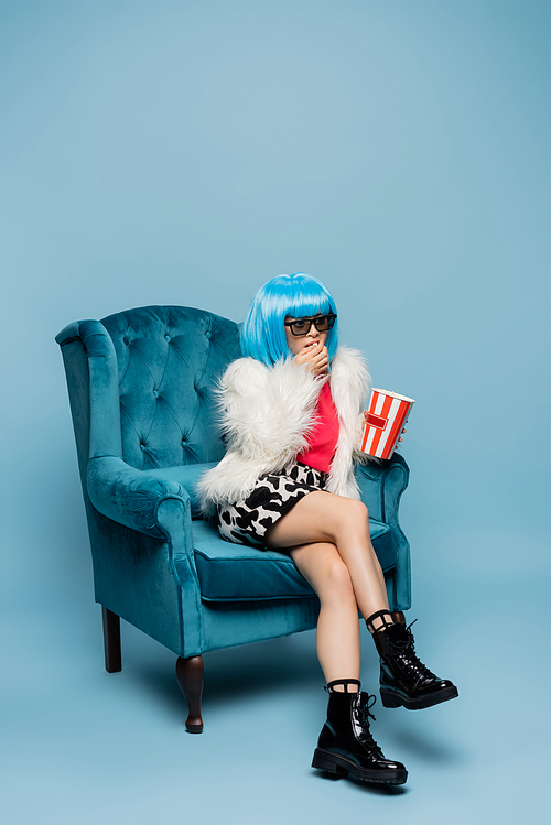 Stylish asian pop art woman in sunglasses eating popcorn in armchair on blue background