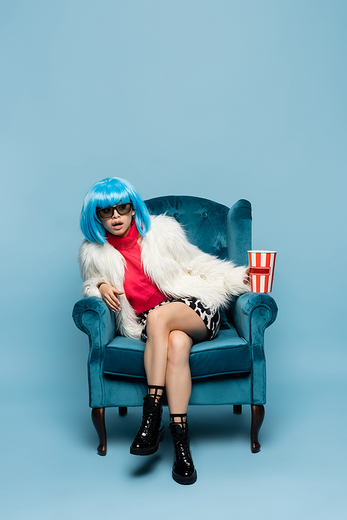 Amazed asian pop art woman in sunglasses holding popcorn on armchair on blue background