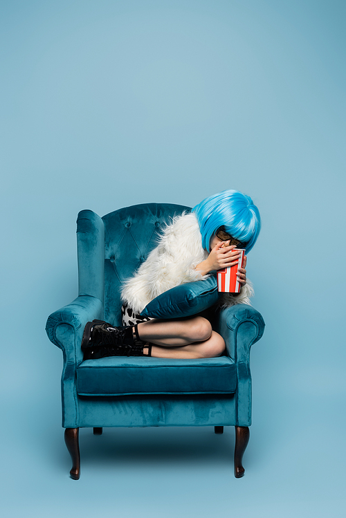 Scared asian pop art woman in bright wig holding popcorn on armchair on blue background