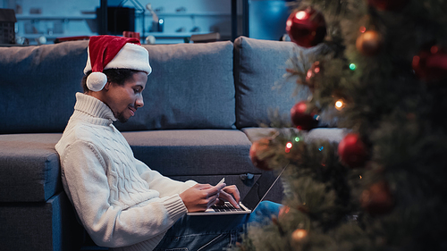 side view of african american man in santa hat using laptop near blurred christmas tree