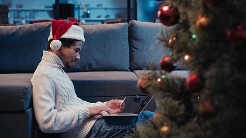 side view of african american freelancer in santa hat using laptop near blurred christmas tree