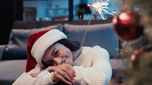 pleased african american woman in santa hat holding sparkler near blurred christmas tree