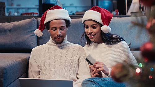 happy african american couple in santa hats online shopping near blurred christmas tree