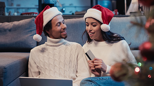 cheerful african american couple in santa hats online shopping near blurred christmas tree